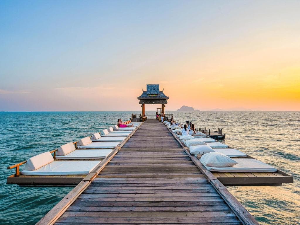 A wooden pier with white cushioned lounge beds extends over the sea toward a small pavilion, with people relaxing and the sun setting in the colorful sky.