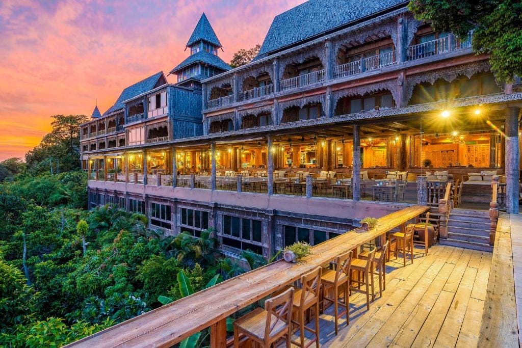 A large, wooden, multi-story building with ornate architecture is lit warmly at sunset. The structure overlooks lush green foliage, with a long wooden terrace lined with chairs and tables in the foreground.