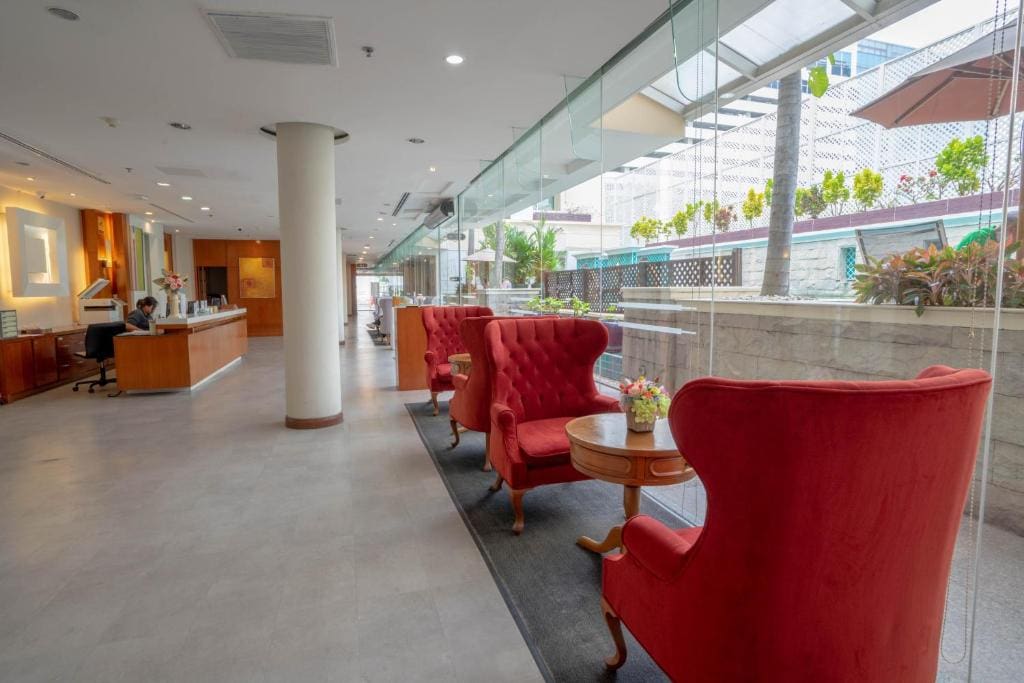 A modern hotel lobby with red armchairs around small tables, large glass windows, potted plants, and a reception desk where a person is seated. Natural light brightens the clean, spacious area.
