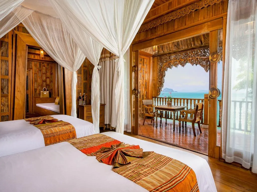 A luxurious wooden hotel room with two beds, white canopies, orange and gold bedding, and a private balcony overlooking the sea. The balcony has a table and chairs, framed by ornate wood carvings.
