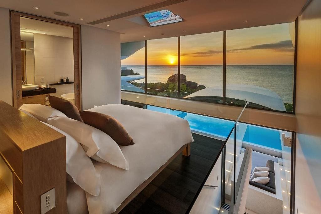 A modern bedroom with floor-to-ceiling windows overlooks an infinity pool and ocean at sunset. The room features a large bed with multiple pillows and an open bathroom visible through a glass wall.