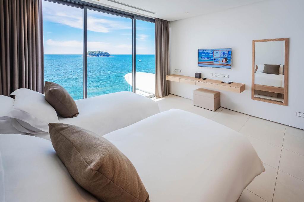 A modern hotel room with two beds, large windows offering an ocean view, a wall-mounted TV, a mirror, and minimalistic decor. Sunlight brightens the white, spacious room, and blue water is visible outside.