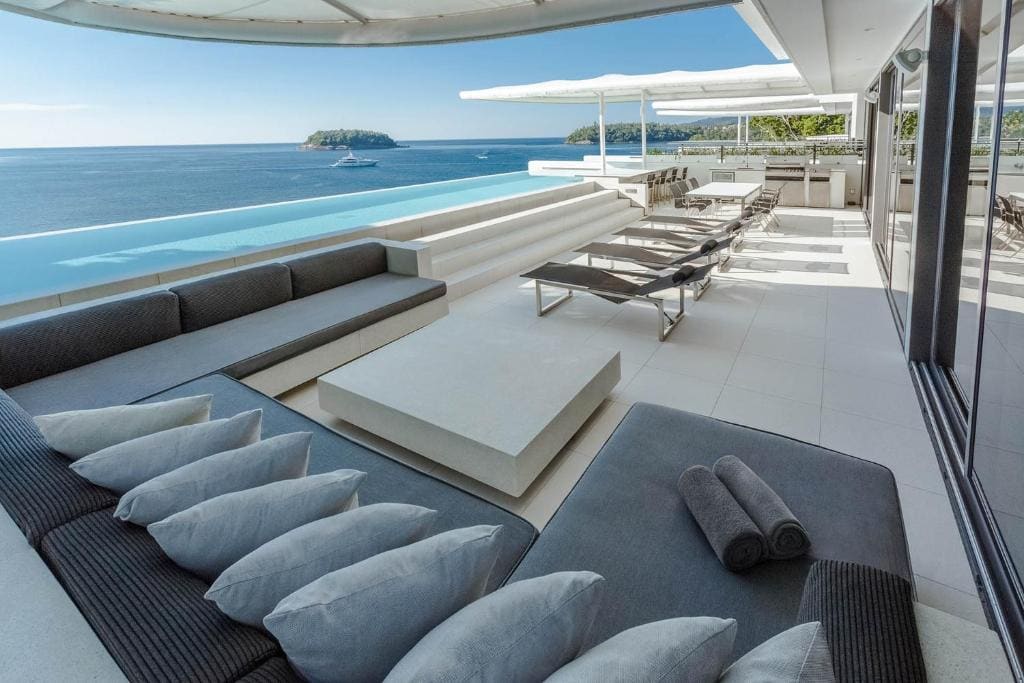 Modern outdoor terrace with cushioned seating, sun loungers, and an infinity pool overlooking the ocean. The scene is bright and spacious, with a clear view of the sea and a small island in the distance.