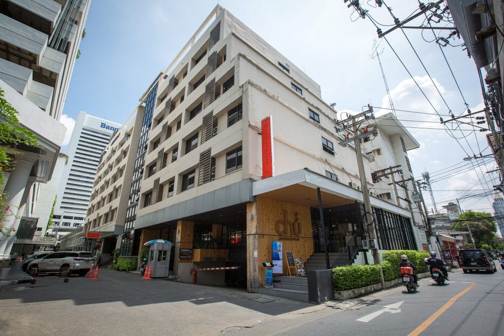 A modern, multi-story building on a city street corner, with a wooden entrance and red signage. Motorbikes and cars are parked nearby, and power lines are visible above. Other tall buildings are in the background.