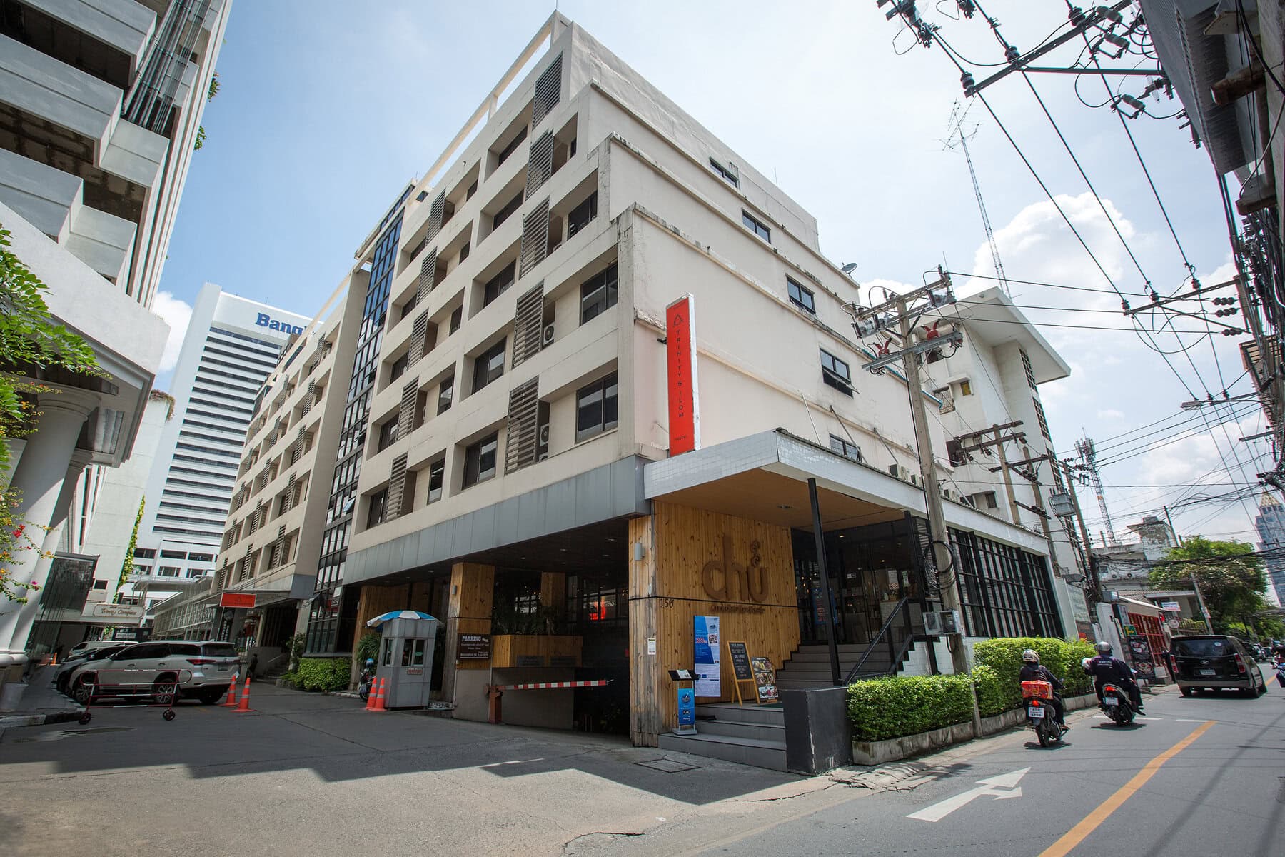 A modern, multi-story building on a city street corner, with a wooden entrance and red signage. Motorbikes and cars are parked nearby, and power lines are visible above. Other tall buildings are in the background.