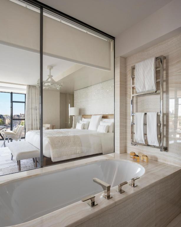 A modern hotel room with a large bed, white linens, and a glass wall separating the bedroom from a bathroom with a sleek bathtub and towel rack. Natural light fills the room through floor-to-ceiling windows.