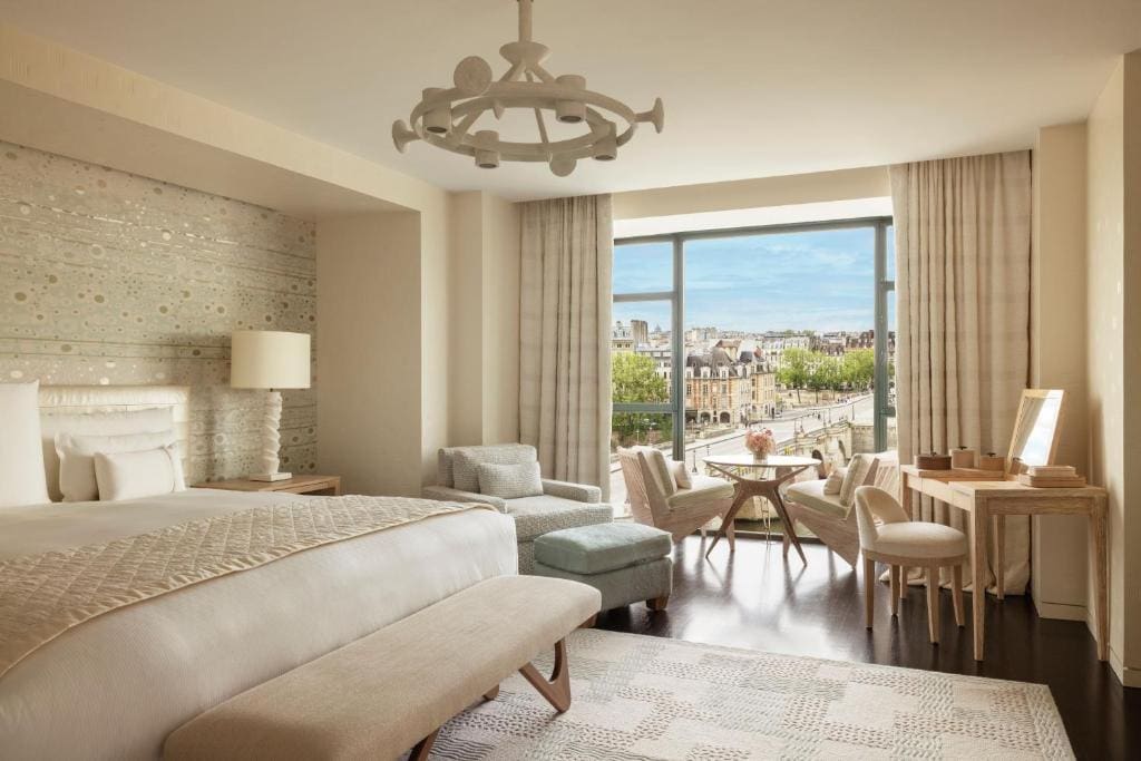 A bright, elegant hotel room with a large bed, seating area by a window, small round table, and a desk. Floor-to-ceiling windows offer a scenic city and river view. Light, neutral tones decorate the space.
