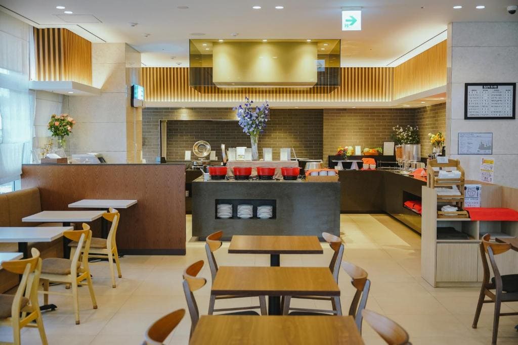 A modern, clean cafeteria with wooden tables and chairs, a buffet-style counter with food trays, and shelves with stacked plates under warm lighting. The space has a cozy and organized atmosphere.