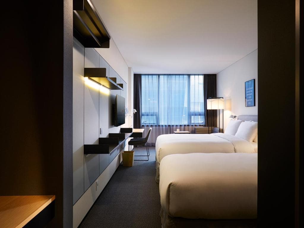 Modern hotel room with two large beds, a desk and chair by a window with sheer curtains, a wall-mounted TV, and minimalist decor in neutral tones. Ambient lighting creates a warm, inviting atmosphere.