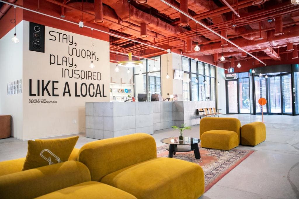 Modern lounge area with mustard-yellow sofas, a glass coffee table, and a reception desk. The ceiling is painted red, and a wall sign reads “Stay, Work, Play, Inspired Like a Local.” Large windows bring in natural light.
