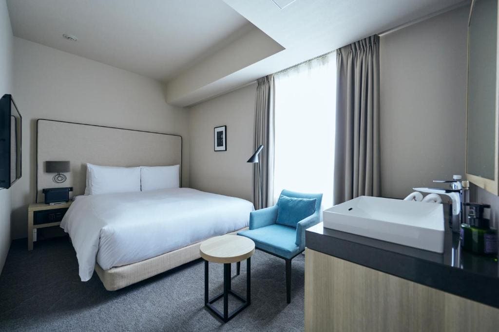 A modern hotel room with a large bed, a blue armchair, a small round table, a wall-mounted TV, a sink with toiletries on a countertop, and a large window covered by curtains letting in natural light.