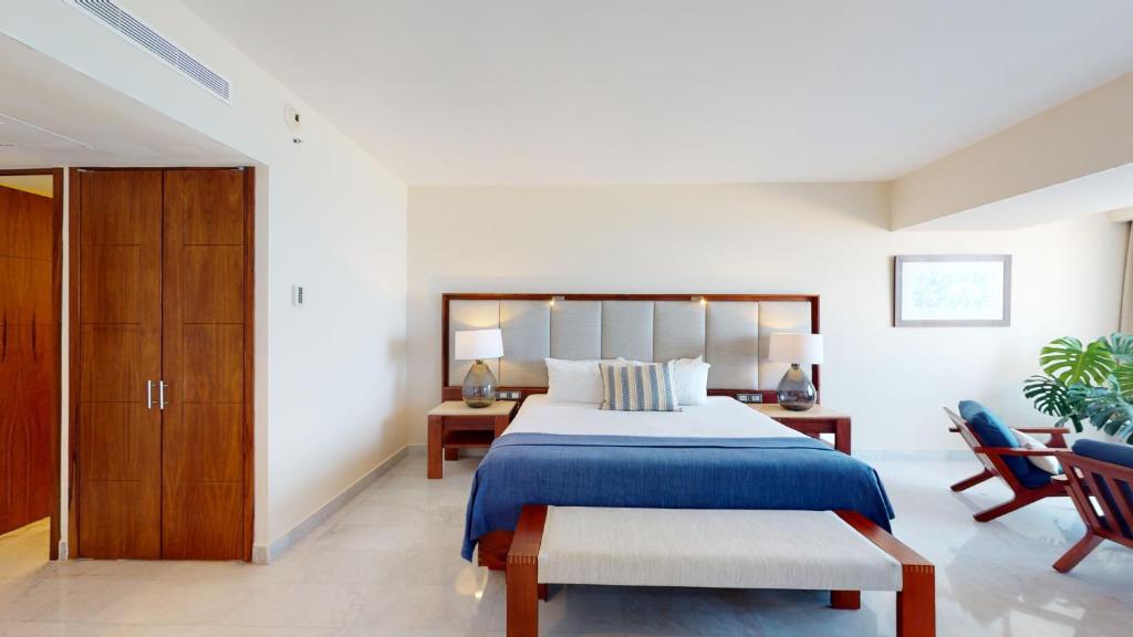 A modern bedroom with a large bed featuring a blue blanket and striped pillows, two bedside tables with lamps, a wooden bench at the foot of the bed, and a lounge chair near a window with natural light.