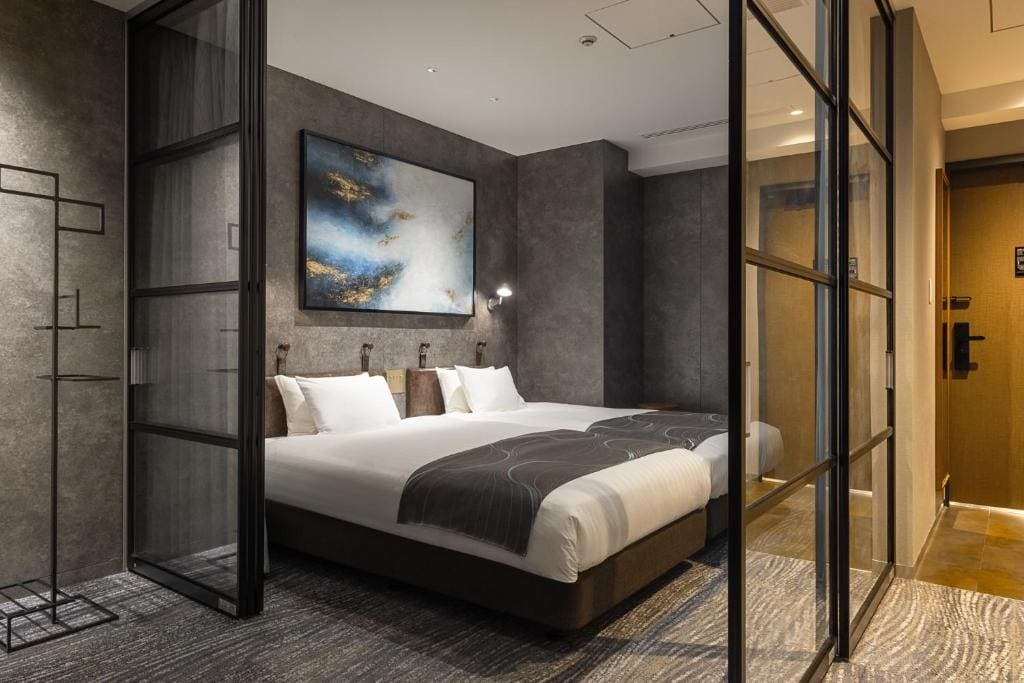 Modern hotel room with two twin beds pushed together, gray accent walls, abstract artwork above the bed, glass partition doors, and contemporary lighting. The room has a clean, minimalist design and a cozy atmosphere.