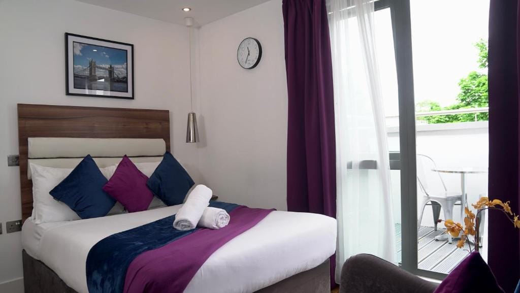 A modern hotel room with a double bed featuring white, blue, and purple bedding, pillows, and a rolled towel. There is a photo above the bed, a clock on the wall, and a balcony with a table and chair outside glass doors.