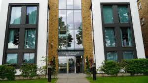 Front view of a modern brick and white apartment building with large windows, green curtains, and a glass entrance labeled Camden Prime Apart Hotel. There are bushes and a pathway in front.