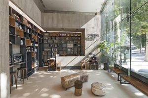 Modern coffee shop interior with a concrete counter, shelves filled with products, stools, soft seating, large glass windows, potted plant, and natural light streaming in from outside.