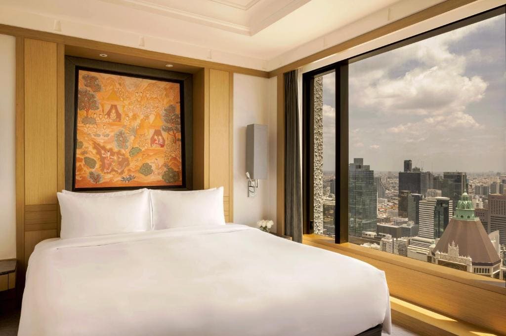 A modern hotel room with a large bed, white bedding, a colorful painting above the headboard, and a floor-to-ceiling window offering a city skyline view under a partly cloudy sky.