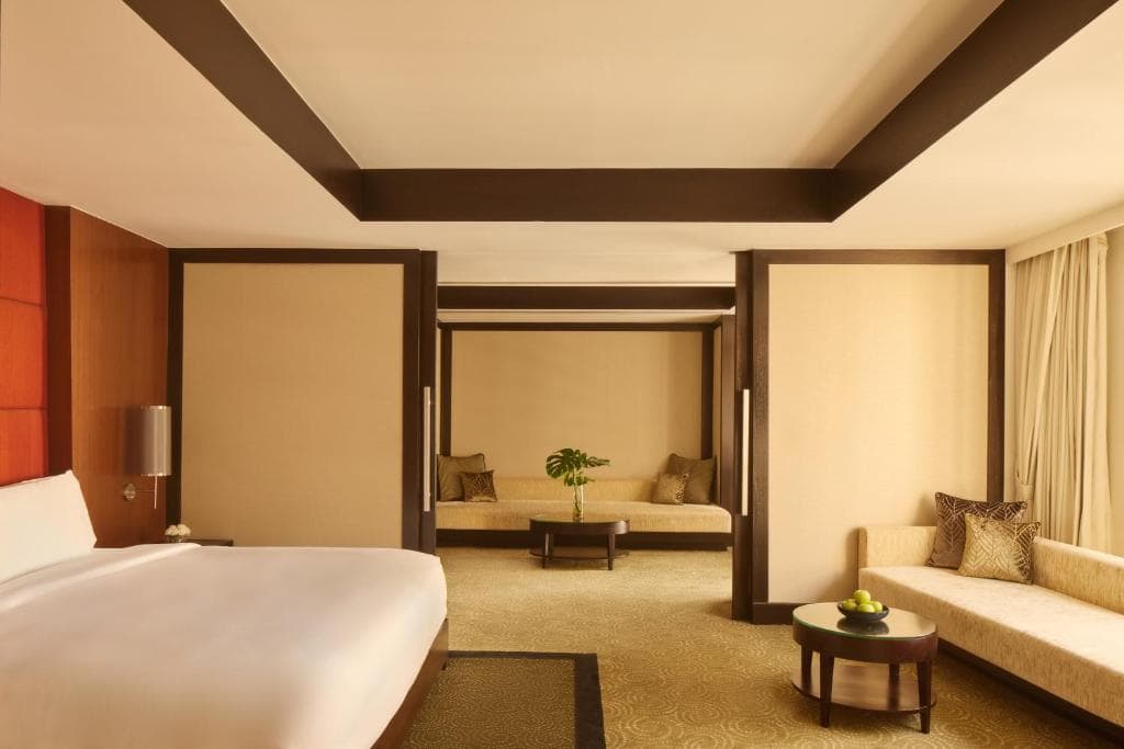 Spacious hotel room with a large bed, two cushioned bench seats, a round coffee table with green apples, beige walls, carpeted floor, and floor-to-ceiling windows with light curtains.