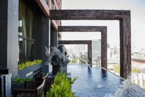 An infinity pool with a decorative fish statue and water spouts on a modern building’s terrace, overlooking a cityscape with tall buildings in the background.