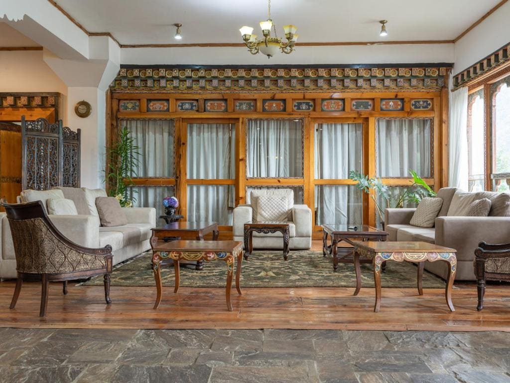 A spacious living room with upholstered sofas, armchairs, ornate wooden tables, large windows with curtains, indoor plants, and a chandelier hanging from the ceiling. The decor features intricate patterns and wood accents.