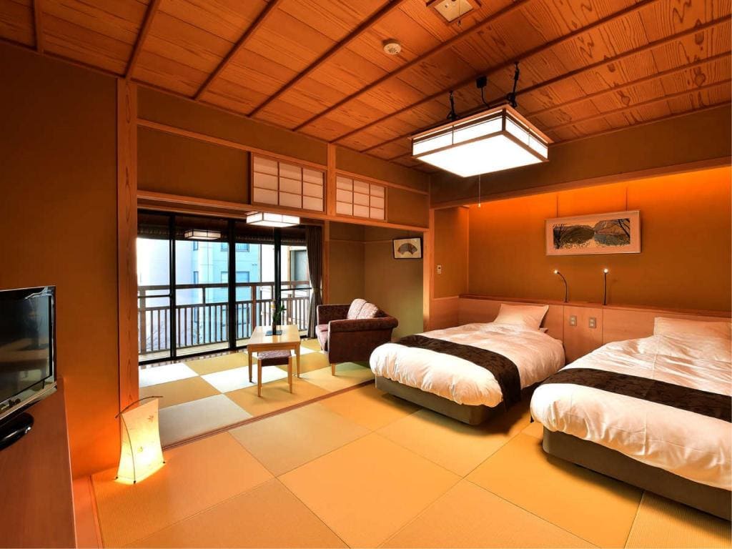 A traditional Japanese-style room with tatami mats, two beds with white bedding, a small table with floor chairs, a balcony with a view, and wooden ceiling and walls. Soft lighting creates a warm, inviting atmosphere.