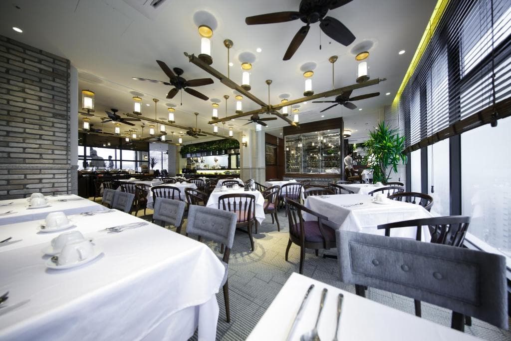 A bright, modern restaurant dining area with white tablecloths, wooden chairs, ceiling fans, and large windows letting in natural light. The tables are set and a person stands near the bar in the background.