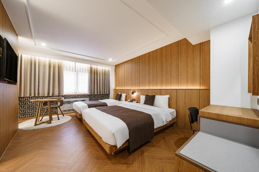 Modern hotel room with two single beds, brown and white decor, wooden floors and walls, a desk, a small table with two chairs by the window, and soft lighting creating a cozy atmosphere.