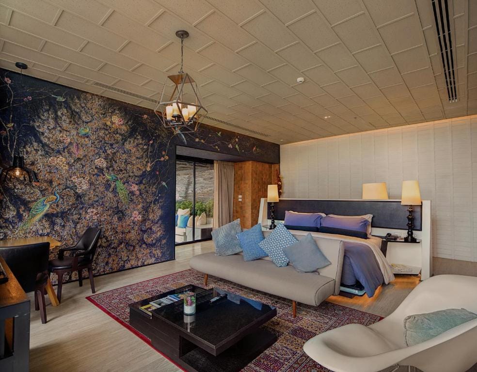 A stylish hotel room with a large bed, blue and white pillows, a patterned rug, a decorative mural wall with peacock designs, modern furniture, and an open doorway leading to an outdoor seating area.