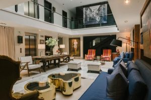 Modern hotel lobby with blue sofas, gold coffee tables, striped armchairs, and art on the walls. The space is open with a mezzanine, elevator, large plants, and contemporary decor. Natural light brightens the area.