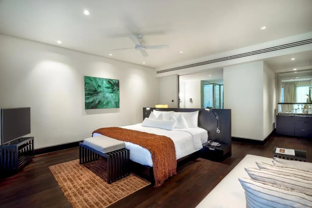 Modern hotel room with a large bed, white bedding, and a brown throw, flanked by nightstands. There is a TV, a bench at the foot of the bed, a green painting on the wall, dark wood floors, and contemporary decor.