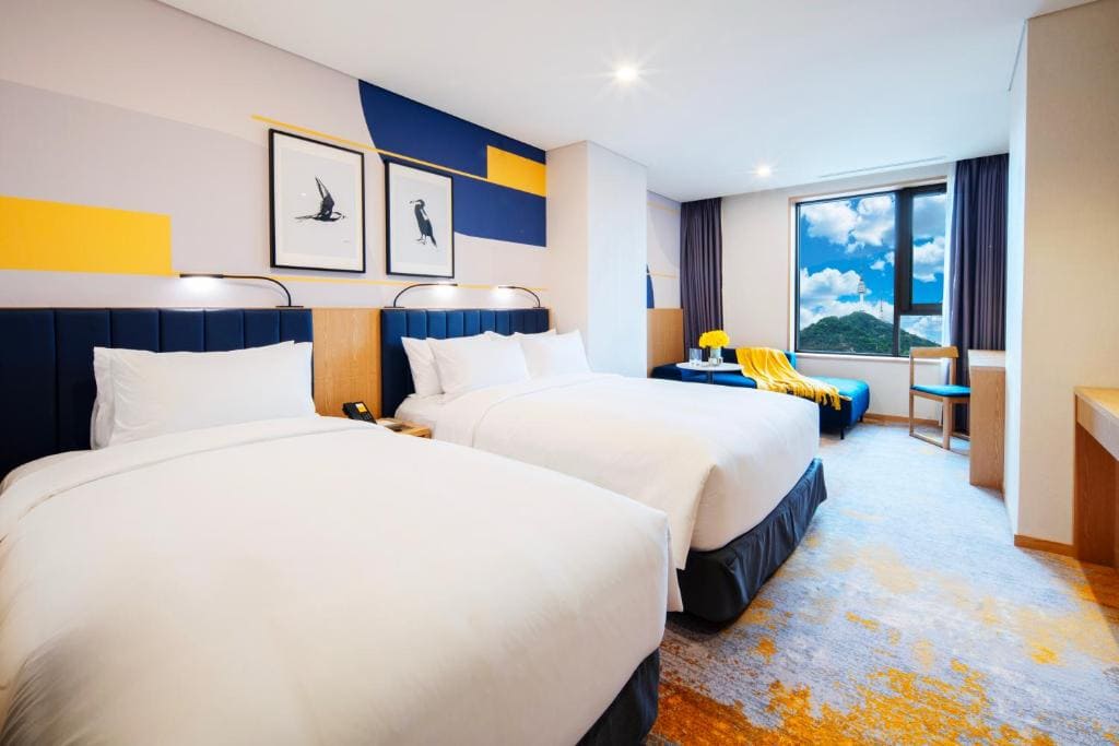 Modern hotel room with two large beds, abstract wall art, a window seat with a yellow blanket, a desk, and a large window showing a scenic view of green hills and a blue sky.