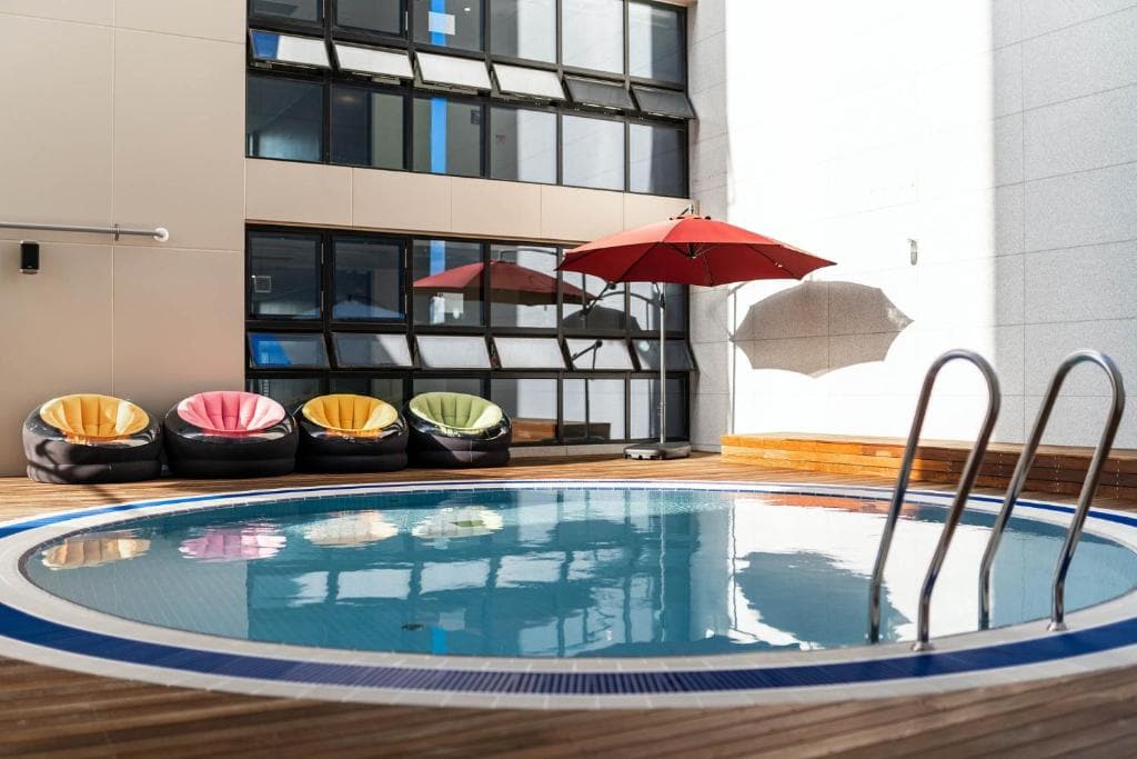 A small round pool on a wooden deck with four colorful bean bag chairs and a red umbrella nearby, set against a modern building with large windows.