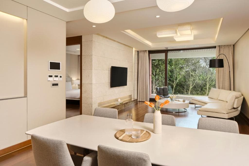 A modern, bright living and dining area with a white table, light grey chairs, a beige sectional sofa, wall-mounted TV, floor-to-ceiling windows, and a view of green trees outside.