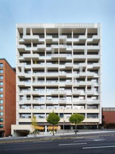 A modern, multi-story apartment building with a white geometric facade, balconies, and vertical railing patterns; a few trees line the sidewalk in front, and adjacent buildings are visible on either side.