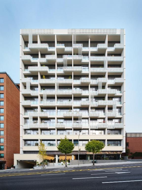 A modern, multi-story apartment building with a white geometric facade, balconies, and vertical railing patterns; a few trees line the sidewalk in front, and adjacent buildings are visible on either side.