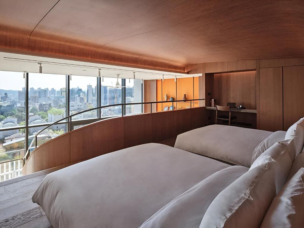 A modern hotel room with two large beds, wood-paneled walls and ceiling, a desk in the corner, and floor-to-ceiling windows offering a city skyline view.