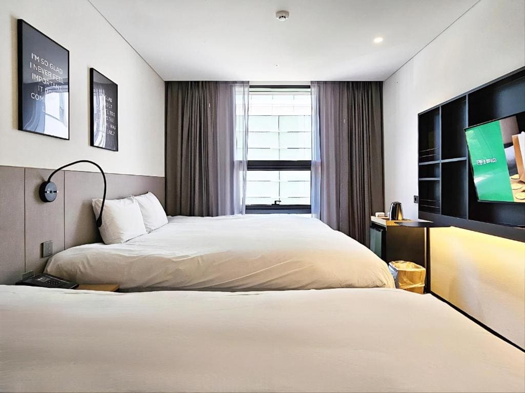 A modern hotel room with two large beds, white bedding, a window with sheer and blackout curtains, wall art, a wall-mounted TV, a shelving unit, and a tea kettle on a small fridge.