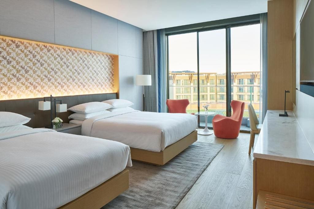 Modern hotel room with two double beds, a decorative headboard, a large window with a balcony view, two coral chairs and a small round table, a desk, and neutral-colored decor.
