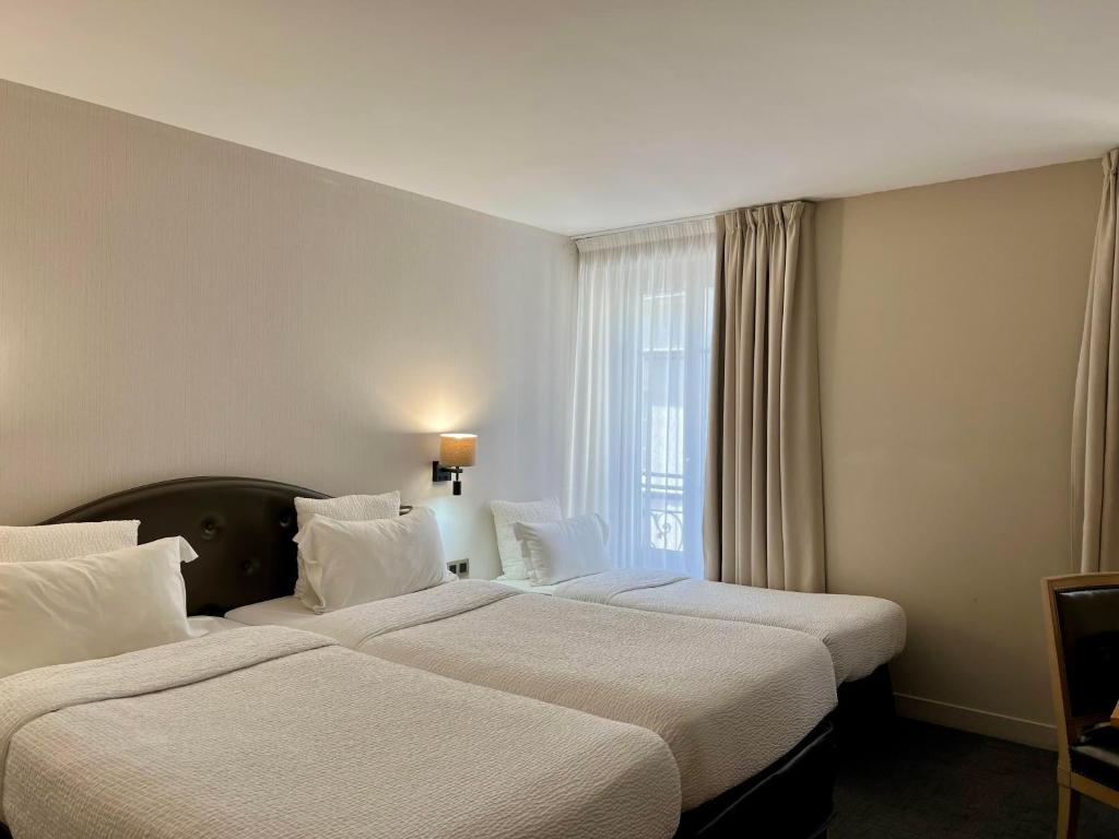 A hotel room with three single beds in a row, each made with white bedding. A wall lamp is above the beds, and natural light enters through a large window with sheer and heavy curtains.
