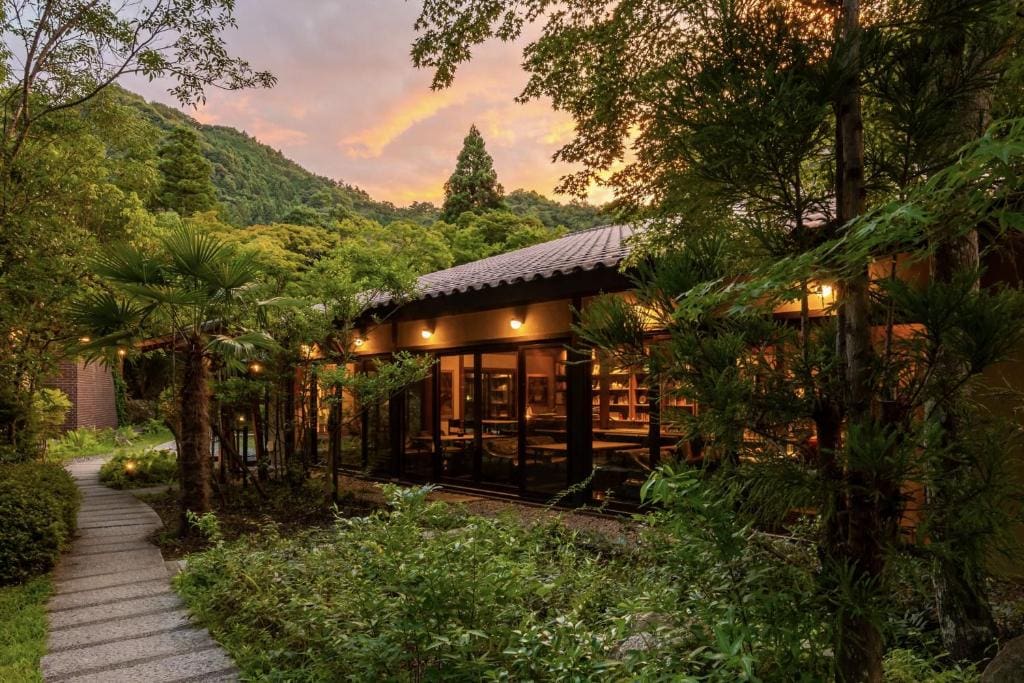 A cozy building with large windows and warm lights sits among lush greenery and trees, with a stone path leading past it. Hills and a colorful sunset sky are visible in the background.