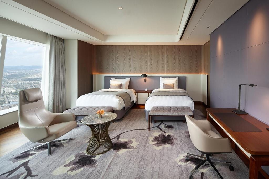 Modern hotel room with two twin beds, a large window with a city view, a lounge chair, a round glass table, and a work desk with a lamp. The room features neutral tones and contemporary decor.
