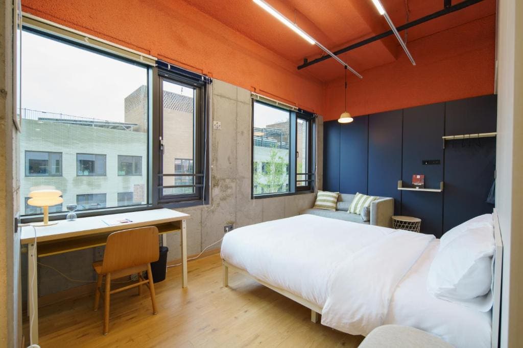 A modern bedroom with large windows, an orange ceiling, a neatly made double bed, a wooden desk and chair, a small sofa with striped cushions, and blue accent walls. Natural light fills the room.