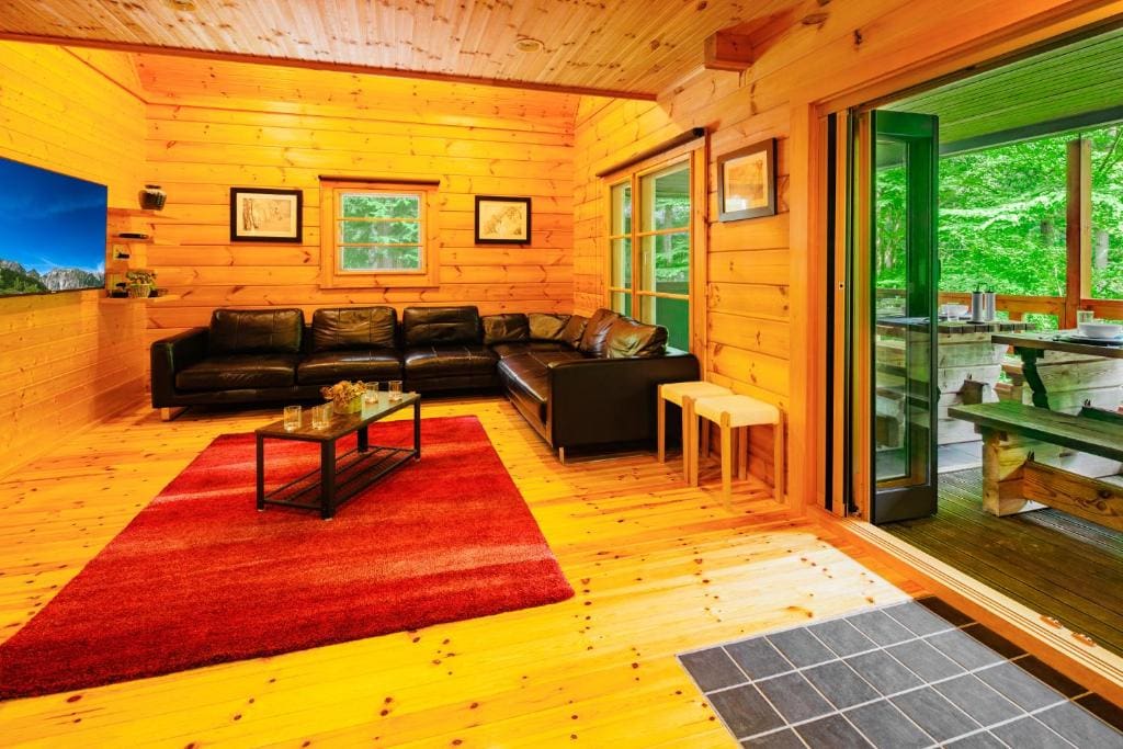 A cozy cabin living room with wood-paneled walls and floors, a black leather sectional sofa, a red rug, wall art, and large glass doors open to a covered deck with a picnic table and forest view.