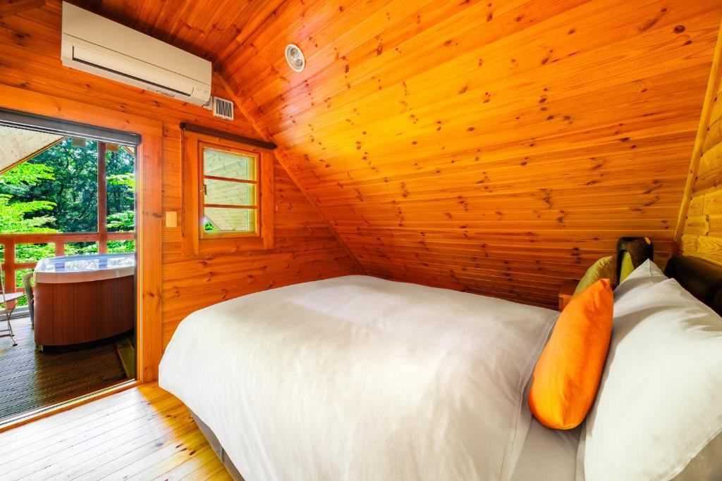 A cozy wooden cabin bedroom with sloped ceiling, white bedding, and orange accent pillows. A door opens to a balcony with a hot tub and a view of green trees outside.