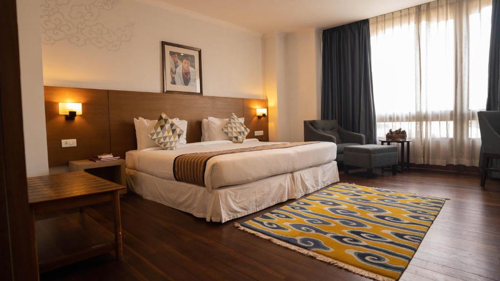 A modern hotel room with a large bed, patterned pillows, wall lamps, a wooden headboard, a colorful rug, two armchairs by a window with sheer curtains, and a small table. Warm lighting creates a cozy atmosphere.