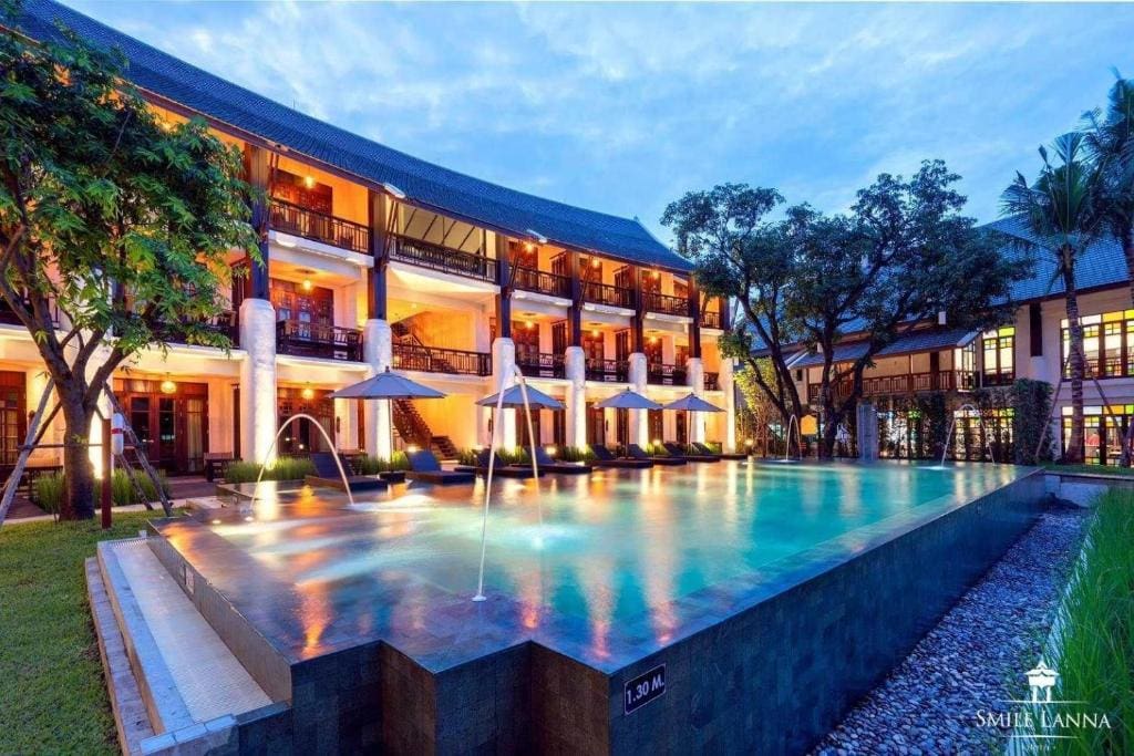 A luxurious hotel with warm lights and balconies overlooks a modern infinity pool surrounded by umbrellas and trees at dusk, creating a serene and inviting atmosphere.