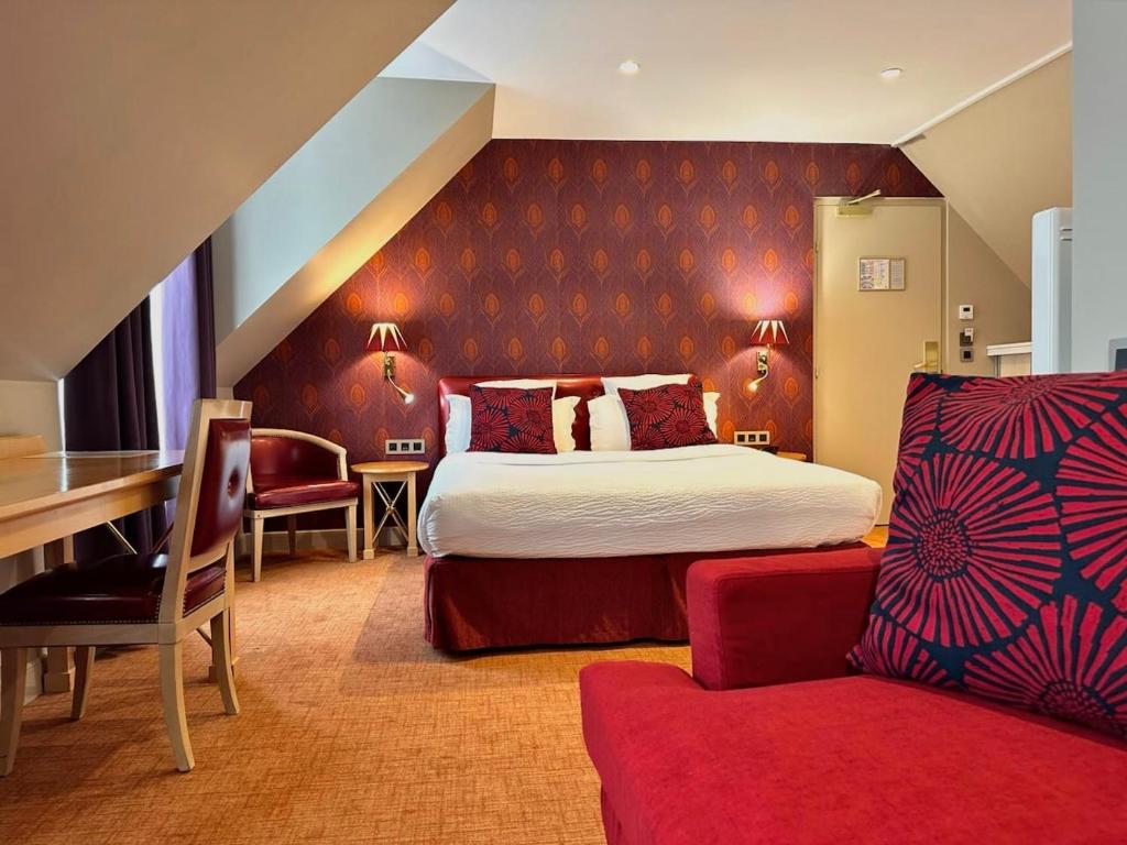 A cozy hotel room with a double bed, accented by red and patterned pillows, red wallpaper, a desk with two chairs, a red armchair, and warm lighting creating an inviting atmosphere.