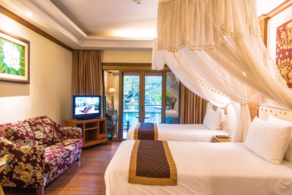 A hotel room with two canopy-covered beds, a floral patterned sofa, a TV on a wooden stand, and a glass door leading to a balcony with outdoor seating and greenery visible outside.