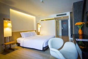 A modern hotel room with a large bed, white linens, two bedside lamps, a desk with a white chair, a vase of orange flowers, and soft, warm lighting.