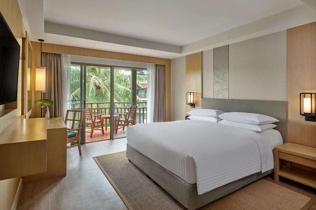 Modern hotel room with a large bed, wooden furniture, TV, and desk. Sliding glass doors open to a balcony with chairs, overlooking palm trees and greenery outside. Soft, natural light fills the room.